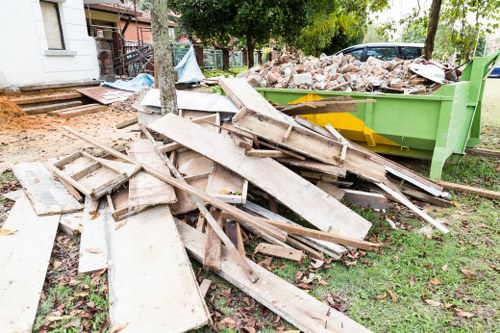 A pile of wooden and concrete debris, including weathered planks, broken boards, and fragments of brick, is scattered on a grassy area outside a residential property. The materials are stacked in an unorganized manner, with some leaning against a green skip filled with more rubble and broken bricks, situated on a paved driveway. In the background, there are trees with green foliage, a white building with steps, and additional construction waste visible. The scene appears to be part of a rubbish removal process, typical of an office or home clearance in Gunnersbury, with weather conditions indicating an overcast day. The presence of the skip and debris suggests professional rubbish removal services by Office Clearance Gunnersbury or similar local providers.