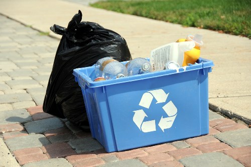 A blue plastic recycling bin with a white recycling symbol on the side, filled with clear plastic bottles and a few other waste containers, placed on a paved driveway next to a black garbage bag. The scene is set outdoors in an area with a grassy verge and a concrete sidewalk, likely in a residential or commercial location within the vicinity of Gunnersbury. The image appears to relate to rubbish disposal or recycling services provided by Office Clearance Gunnersbury, with the bin and waste materials awaiting collection or removal as part of rubbish clearance activities.