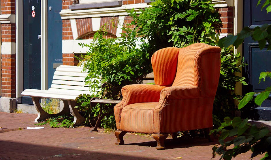 A vintage, upholstered sofa with a dark wooden frame and patterned brown fabric upholstery is positioned directly in front of a large, overflowing wall of compacted waste composed mainly of crumpled c